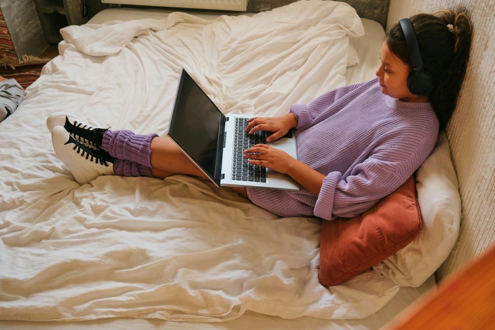 woman shopping online during black friday A person wearing headphones and a purple sweater sits on a bed with white sheets, typing on their laptop to prepare for Black Friday deals.