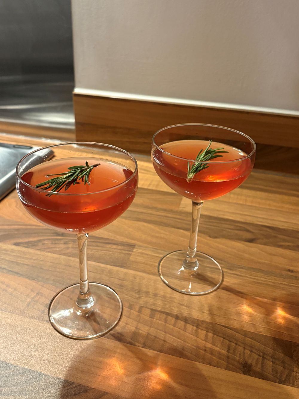 Two coupe glasses filled with a reddish cocktail, each garnished with a sprig of rosemary, sit on a wooden countertop.