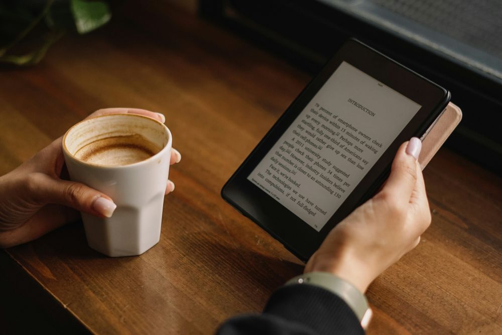 person holding ereader A person holds a white mug of coffee in one hand and an e-reader displaying text in the other, seated at a wooden table by a window.
