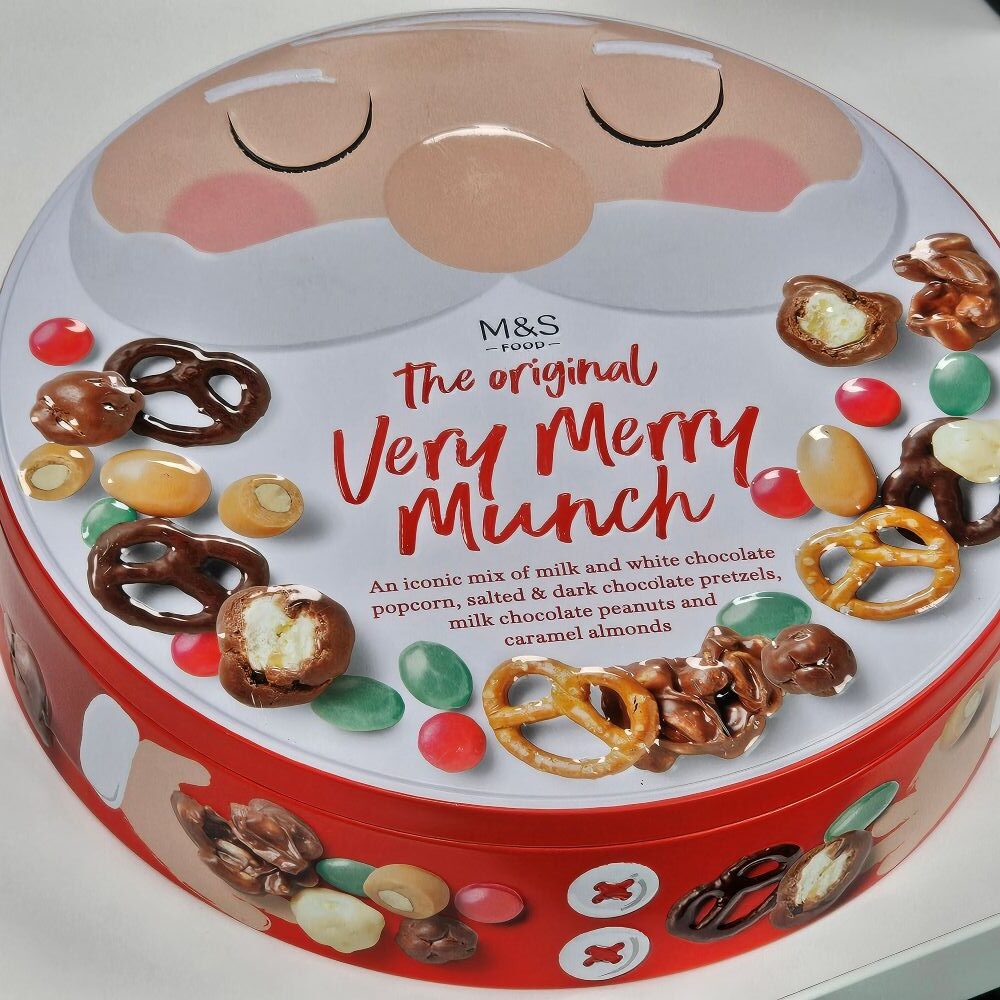tcb marks and spencer the original very merry munch Round tin labeled "M&S Food The original Very Merry Munch" featuring popcorn, pretzels, nuts, and chocolate, with a festive Santa face design on the lid.