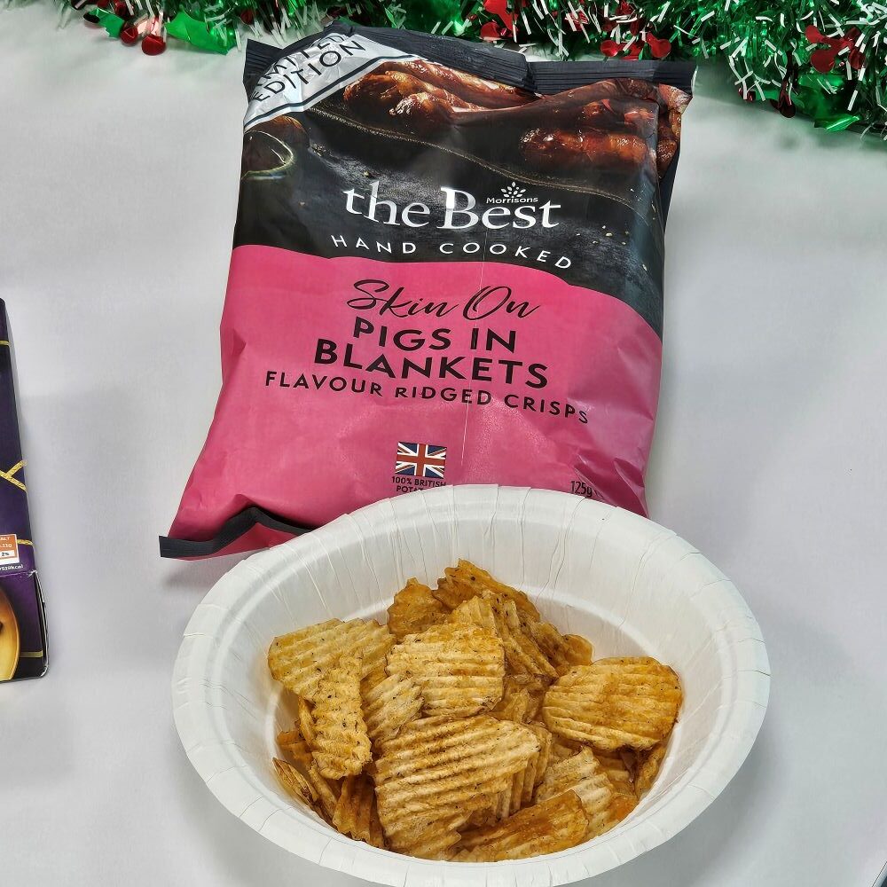 tcb morrisons the best skin on pigs in blankets flavour ridged crisps A paper bowl filled with ridged crisps sits in front of a packet labeled "The Best Hand Cooked Skin On Pigs in Blankets Flavour Ridged Crisps.