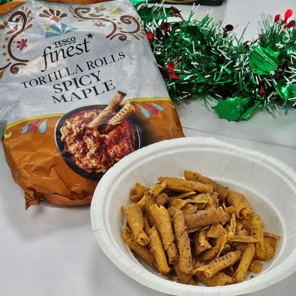 tcb tesco finest maple roll tortilla A paper bowl of Tesco Finest Spicy Maple Tortilla Rolls sits next to the product bag on a white surface with green holiday tinsel in the background.