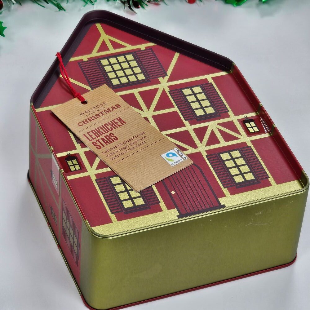 tcb waitrose christmas lebkuchen stars A house-shaped tin with a red and gold design labeled "Finest Christmas Lebkuchen Stars" and a Fairtrade logo, placed on a white surface.