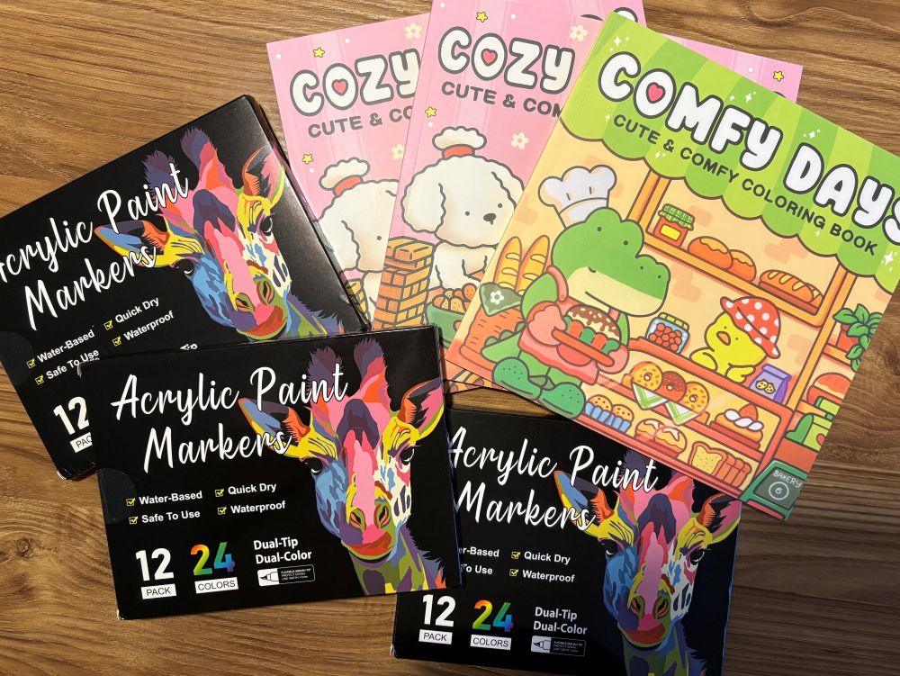 Three boxes of acrylic paint markers and two cute coloring books are arranged on a wooden surface.