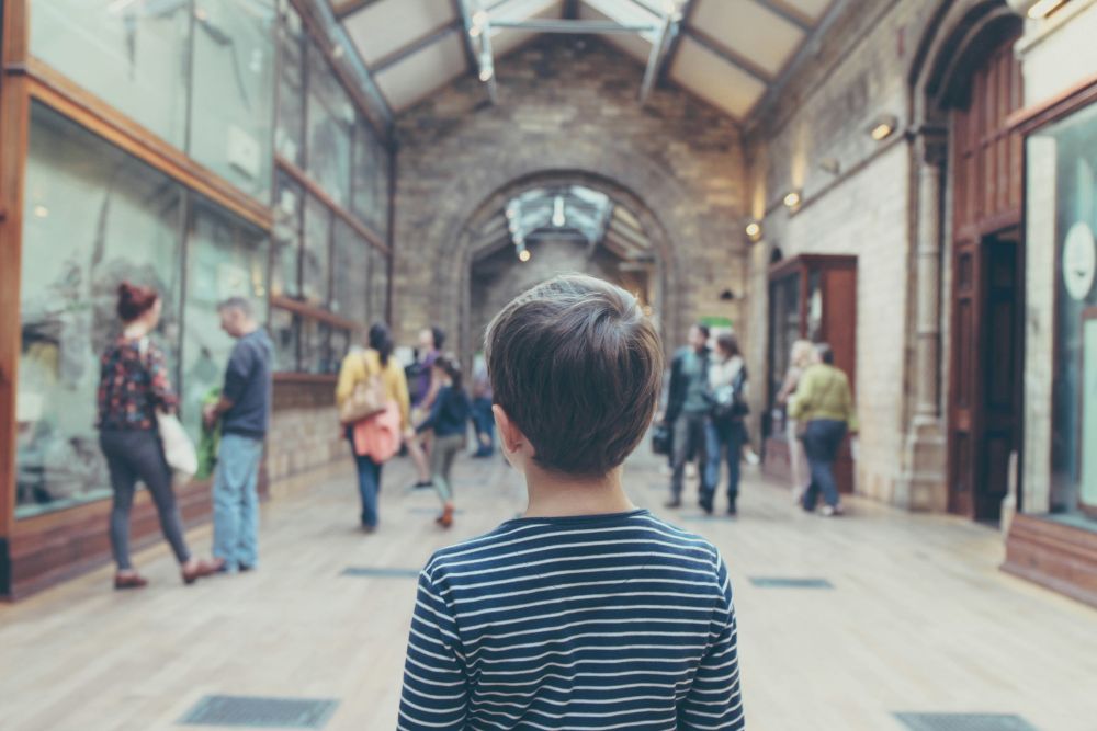 A child in a striped shirt stands in a museum hallway, facing away, while several people look at exhibits and walk around.
