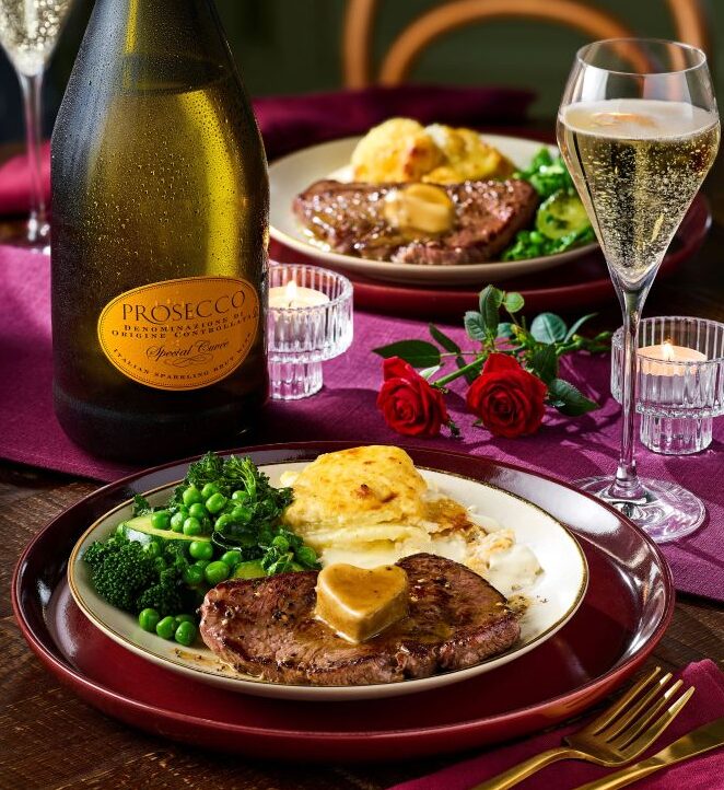 co-op valentines dine in 2026 A dinner table set with steak, vegetables, mashed potatoes, a bottle of Prosecco, two glasses of sparkling wine, candles, and red roses on a wooden surface.