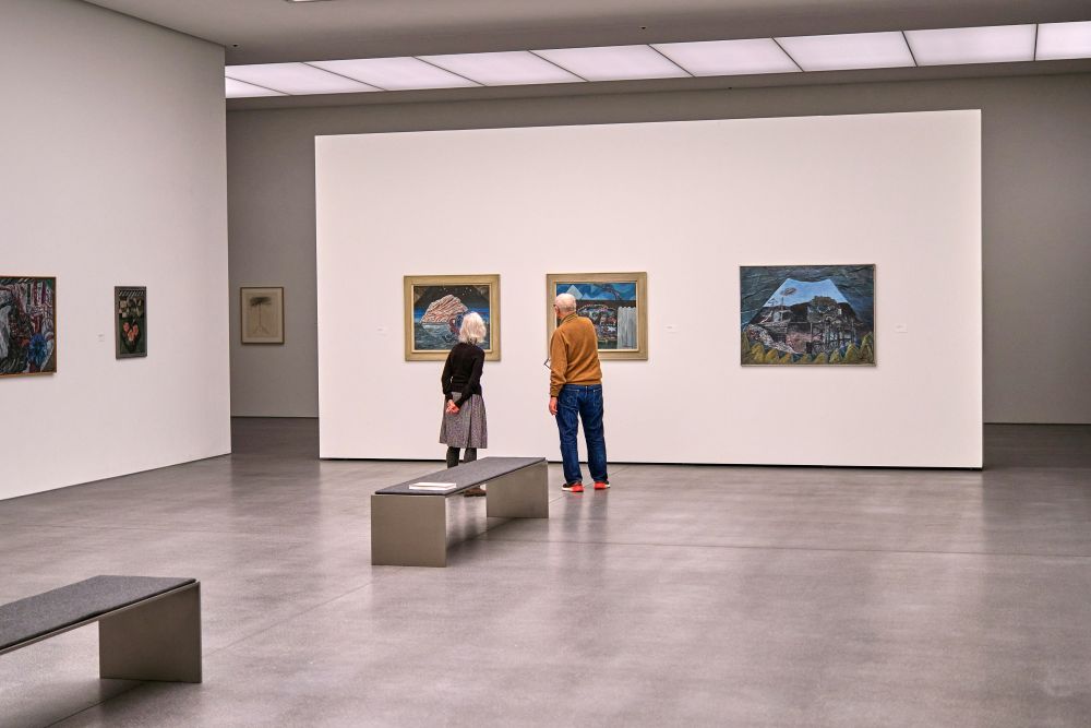 Two people stand in a modern art gallery viewing landscape paintings on a white wall. The room is spacious with benches and additional artwork displayed.