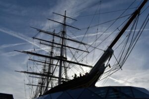 A large historic sailing ship with multiple masts and rigging is silhouetted against the sky, with modern glass structures visible in the foreground.