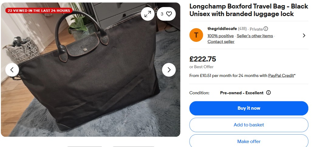 A large black Longchamp Boxford travel bag with brown handles is placed on a grey carpet next to a fluffy rug and a storage basket.