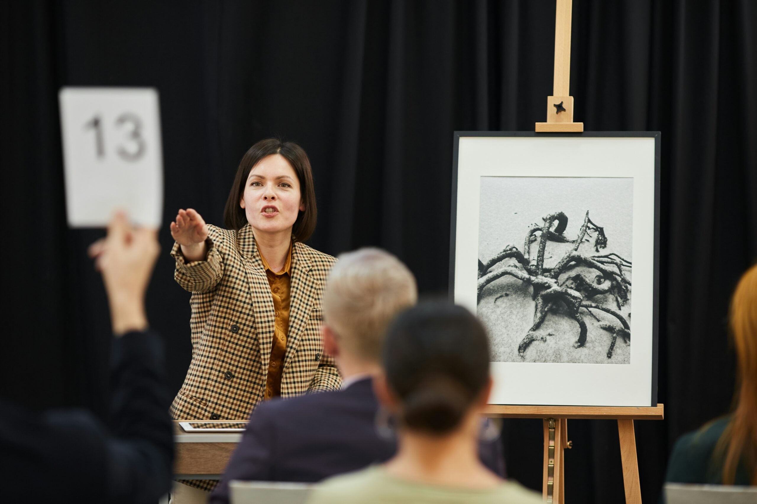 A woman at an auction gestures while someone in the audience raises a card with the number 13; a framed black-and-white artwork of crabs is displayed on an easel nearby.