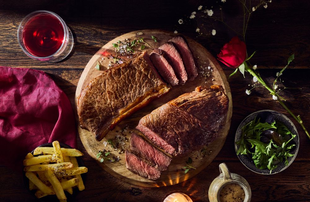 iceland valentines dine in 2026 A sliced cooked steak on a wooden board, surrounded by a glass of red wine, fries, salad, a red napkin, a rose, and a small bowl of sauce on a dark wooden table.