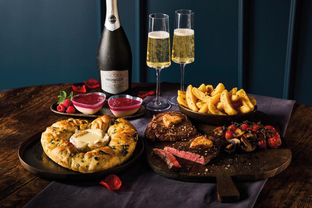 morrisons valentines dine in 2026 A table set with steak, roasted vegetables, cheese bread, potato wedges, two glasses of sparkling wine, a bottle of Prosecco, and two bowls of raspberry dessert.