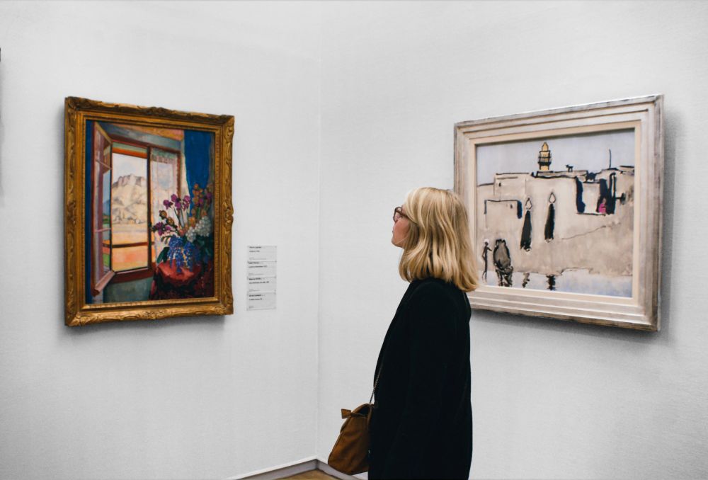 A woman with blonde hair and glasses stands in an art gallery, looking at two framed paintings on a white wall.