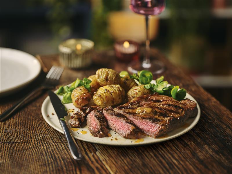 sainsburys valentines dine in 2026 A plate with sliced grilled steak, roasted potatoes, and green salad sits on a wooden table with cutlery and a glass of red wine.