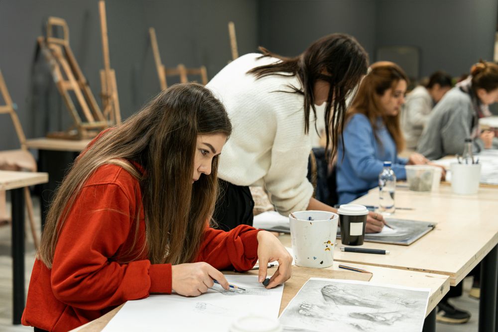 Several people sit at desks drawing on paper in an art classroom, with sketch tools, cups, and easels visible in the background.