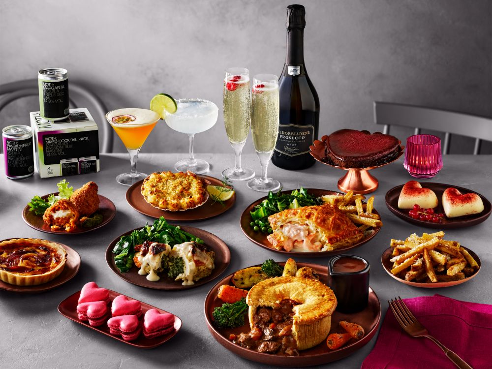 tesco valentines dine in 2026 A table set with various plated dishes, including meat pies, vegetables, desserts, fries, canned cocktails, two cocktails, and a bottle of prosecco with three filled glasses.