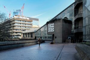 A person walks along a brick pathway outside the Brutalist-style Barbican Centre in London, with construction cranes visible in the background.