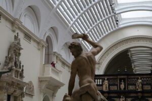A large classical statue of a muscular man holding a raised object stands in a spacious museum hall with arched ceilings and sculptures displayed along the walls.