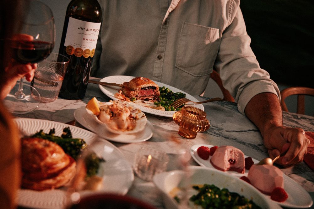 waitrose valentines dine in 2026 A person seated at a marble table with plates of food, wine glasses, a bottle of red wine, and desserts, suggesting a dinner setting.
