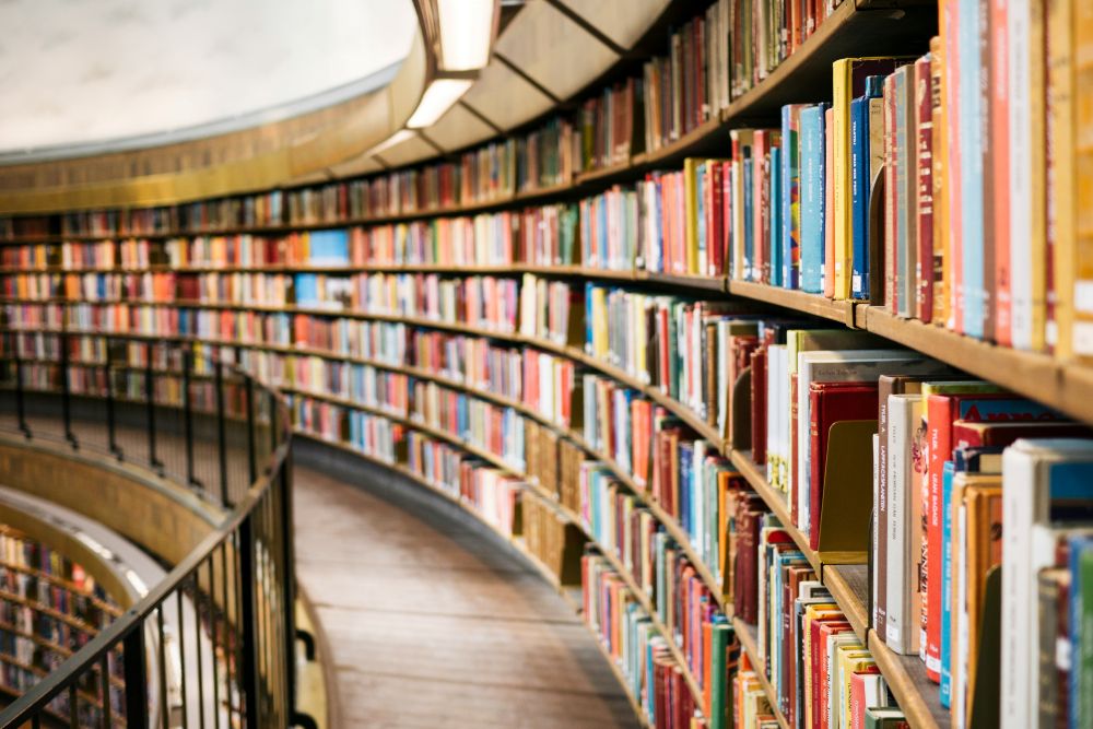 Rows of colorful books line curved wooden shelves in a spacious library, with a walkway and railing along the shelves.
