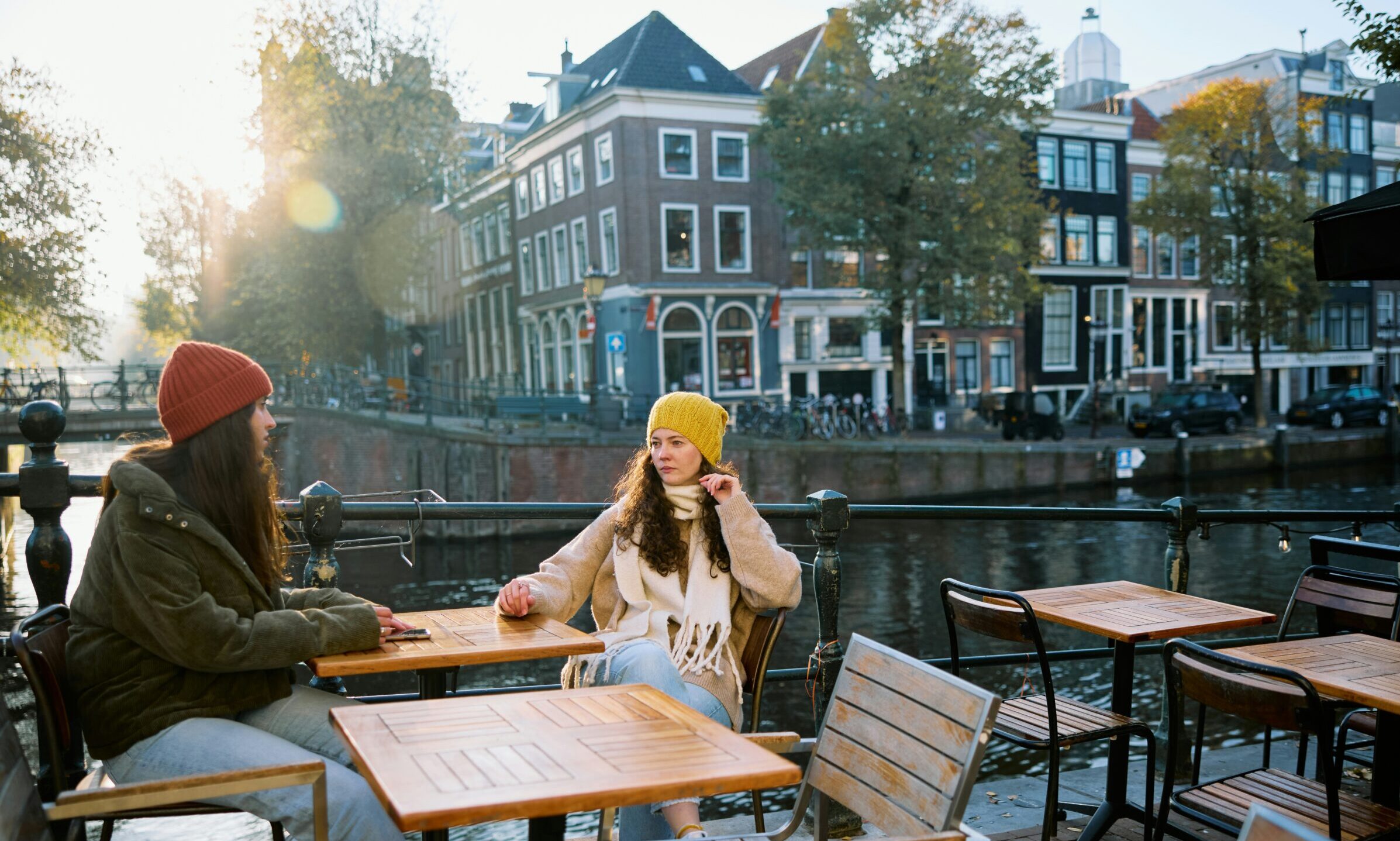eurostar to amsterdam desktop Two people in warm clothing sit at an outdoor café by a canal with traditional European buildings and trees in the background.