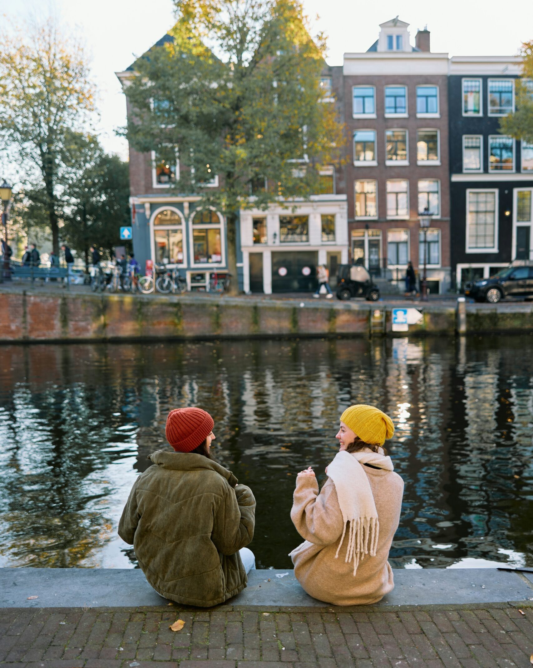 Two people wearing coats and hats sit by a canal, talking. Brick buildings, trees, and bicycles are visible across the water under a clear sky.