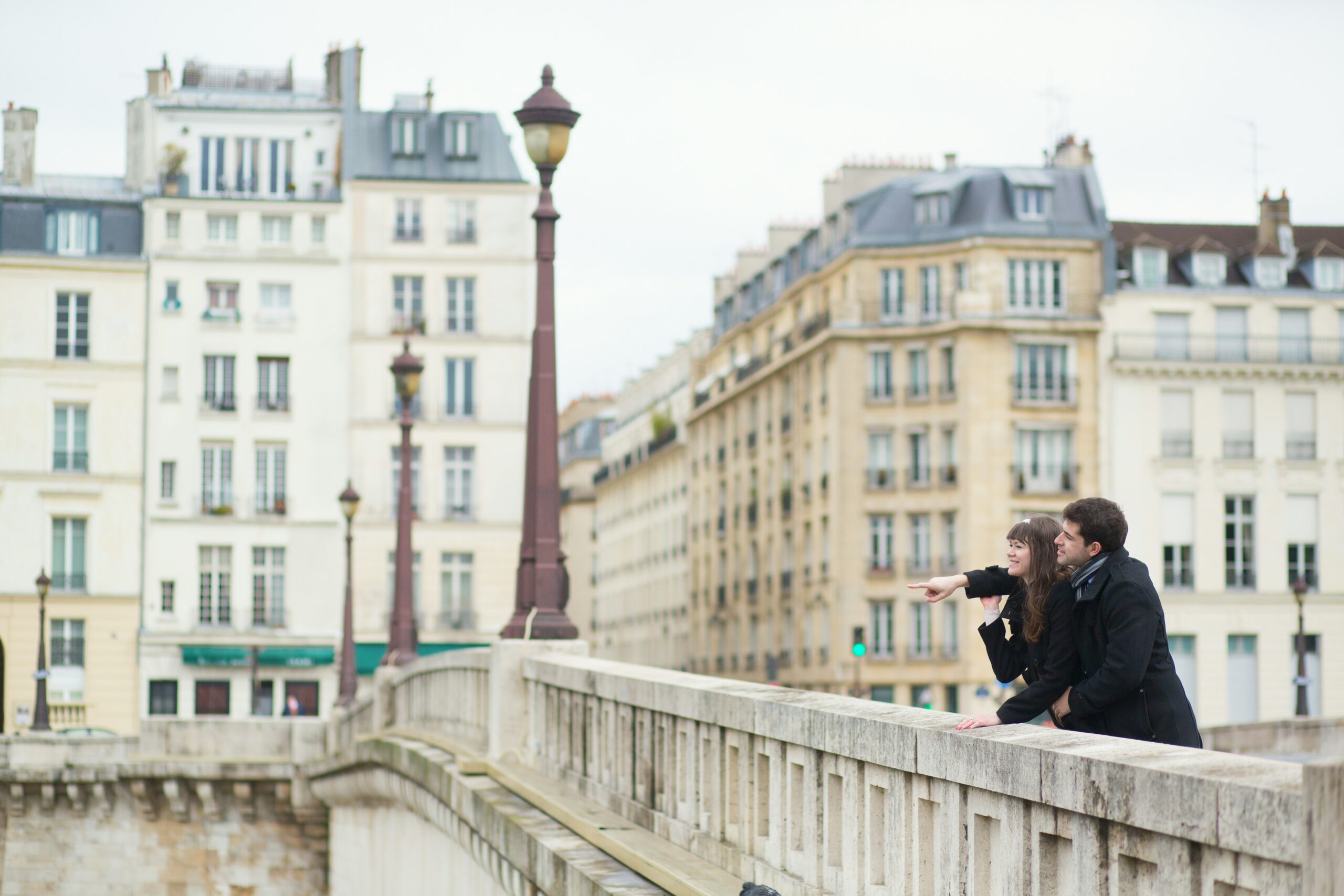 unsplash paris A man and woman stand on a bridge in a city, leaning on the railing as the woman gestures outward. Tall, classic European buildings are visible in the background.