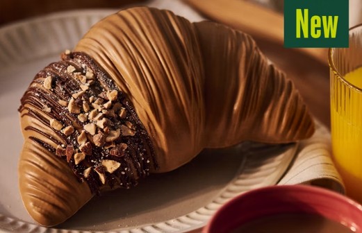 A croissant covered in light brown chocolate and chopped nuts sits on a plate next to a glass of orange juice. A green label in the corner reads "New.
