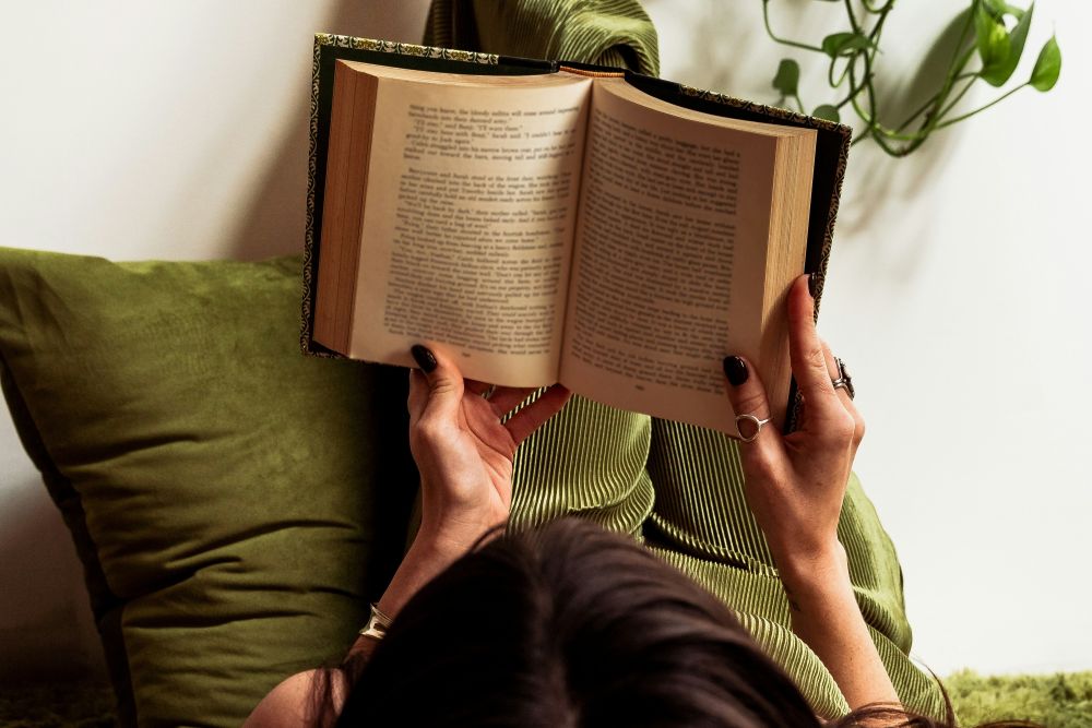 Person lying down on green cushions reading an open book, with hands holding the book and a leafy plant visible in the background.