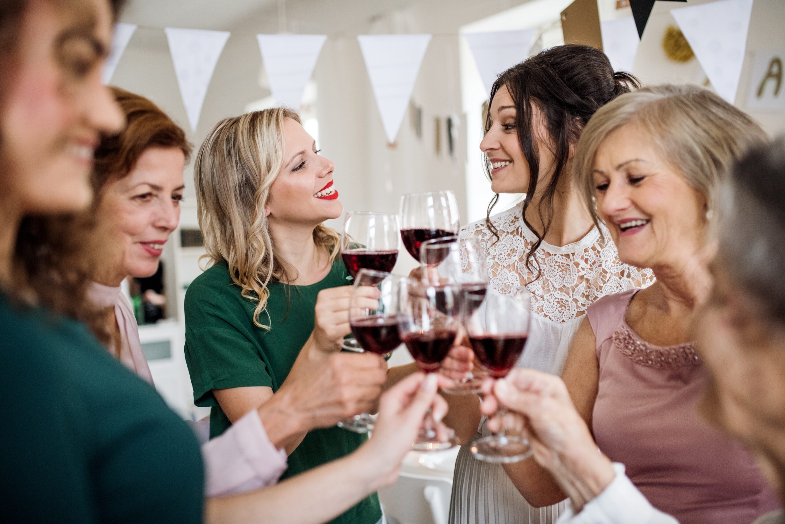getty-images-vi4L-eV9x-M-unsplash A group of women standing in a circle smile and clink glasses of red wine at a celebration, with festive bunting hanging in the background.