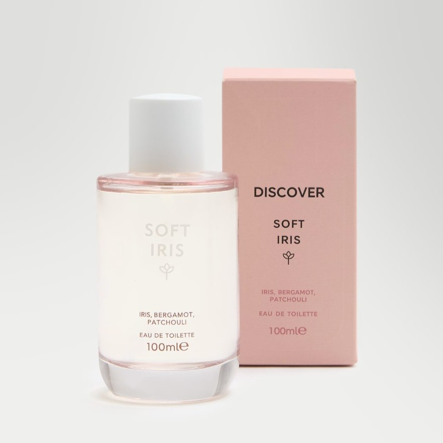 A clear glass bottle of Soft Iris eau de toilette stands next to its pink box packaging, both labeled with scent notes of iris, bergamot, and patchouli, 100 ml size.