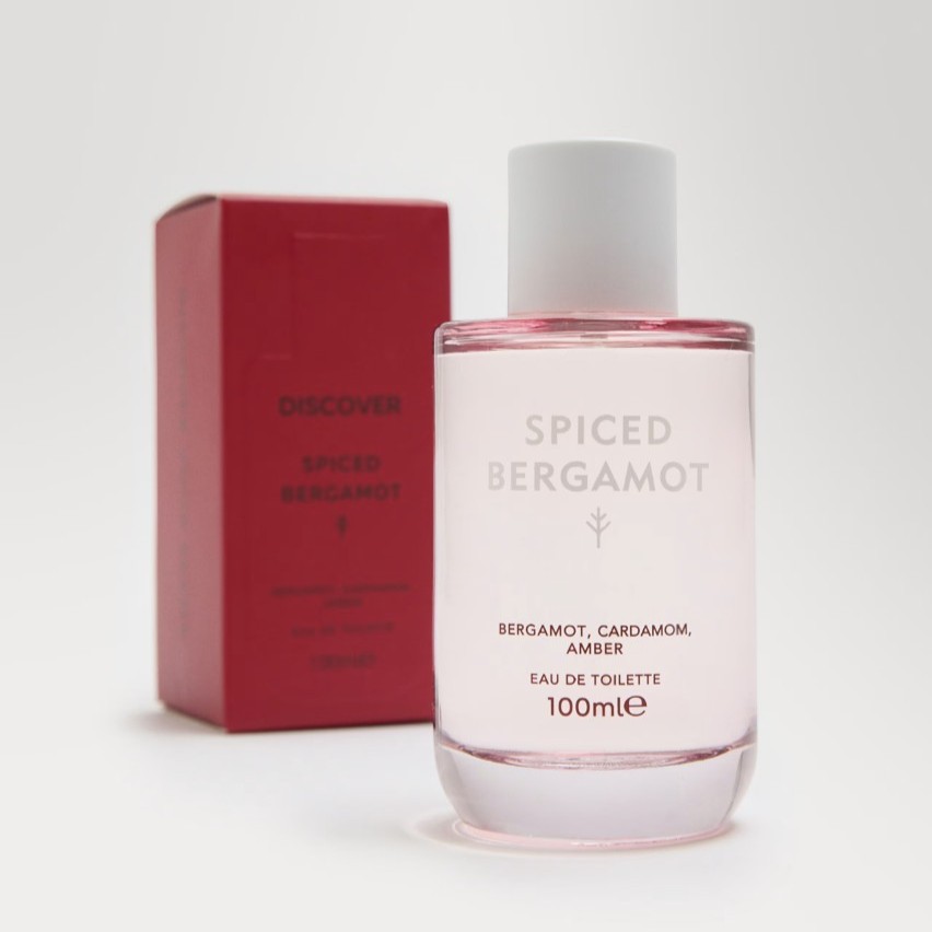 A clear bottle labeled "Spiced Bergamot" Eau de Toilette stands in front of a red box with the same name. The bottle contains 100 ml and lists bergamot, cardamom, and amber.