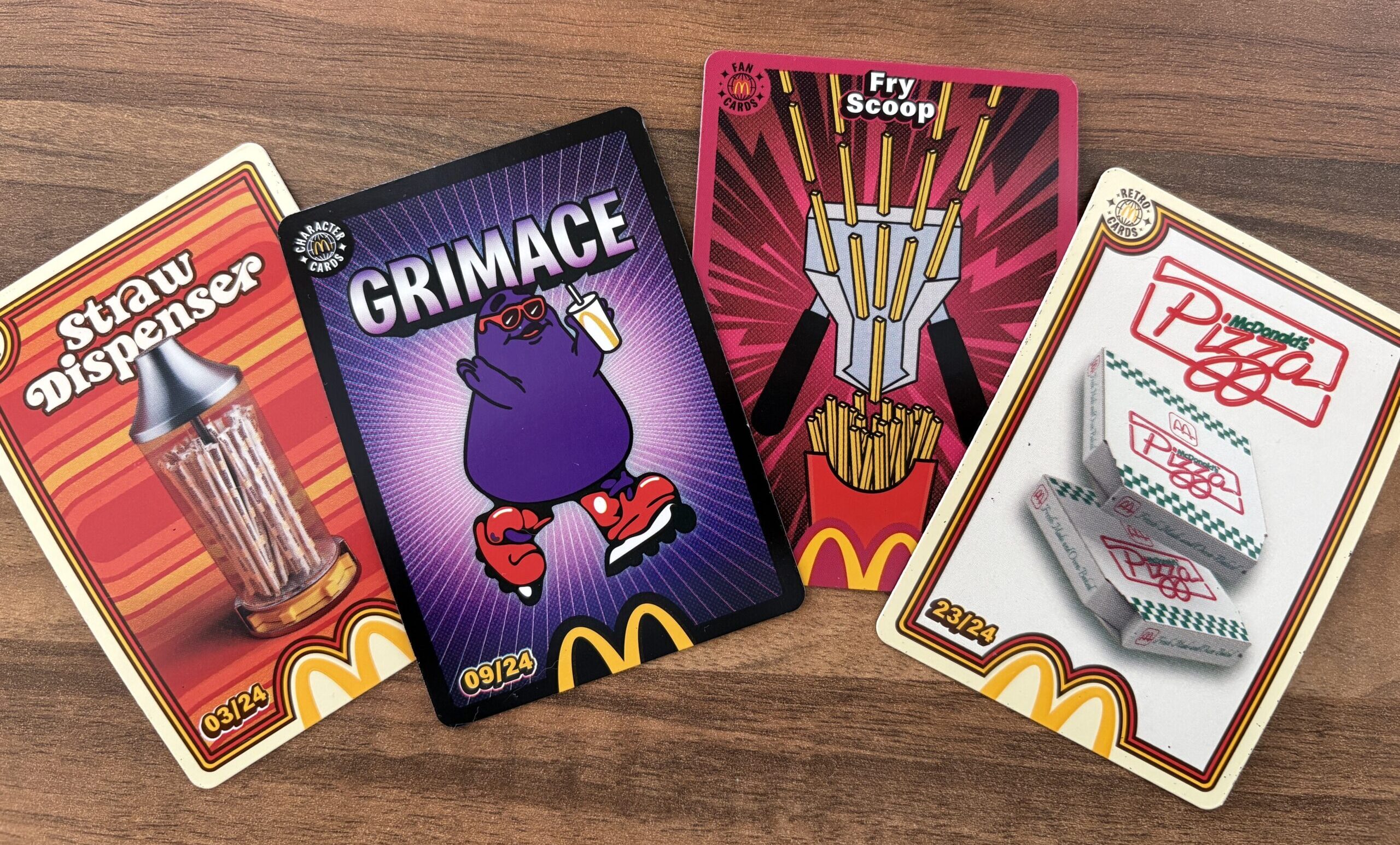 topcashback money guides mcdonalds trading cards Four McDonald’s-themed trading cards are displayed on a wooden surface, featuring images of a straw dispenser, Grimace, a fry scoop, and pizza boxes.