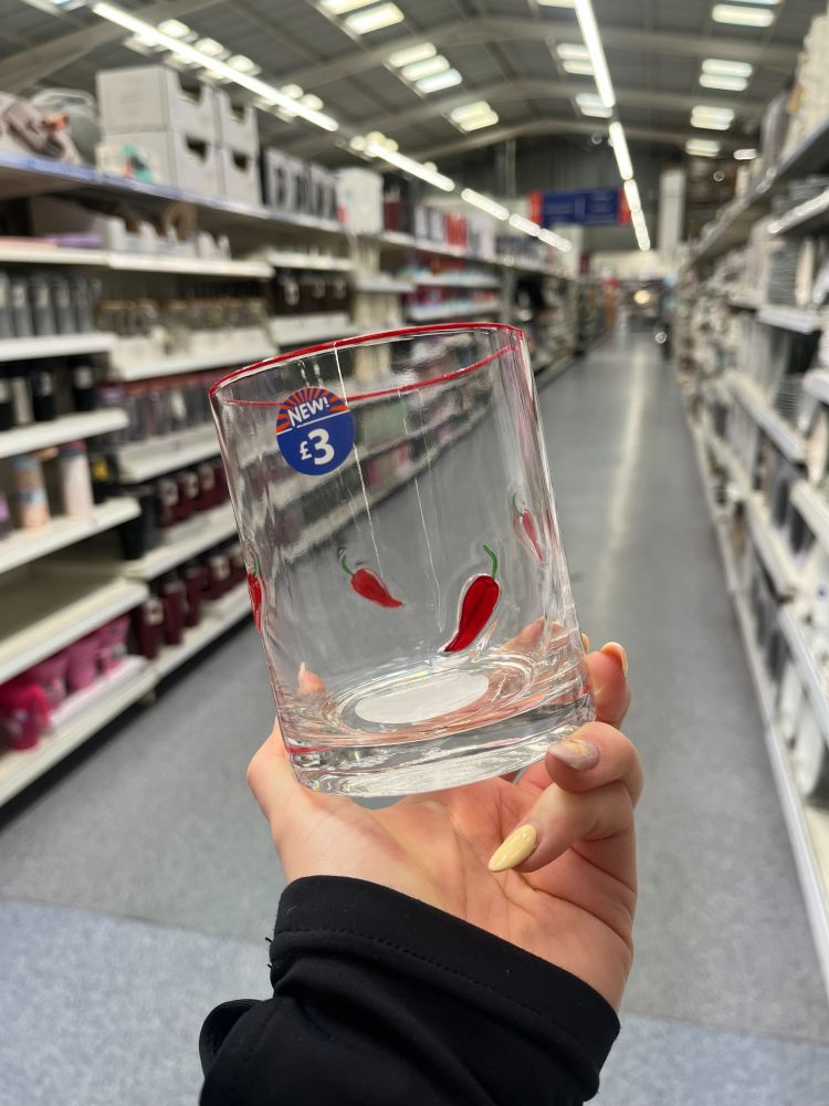 A hand holds a clear glass with red chili pepper designs and a £3 price sticker, inside a brightly lit store aisle.