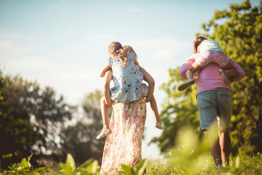 Two adults give piggyback rides to two children while walking outdoors on a sunny day, surrounded by green trees and grass.