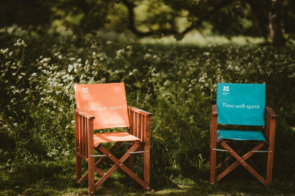 Two wooden National Trust chairs in a garden setting with greenery in the background