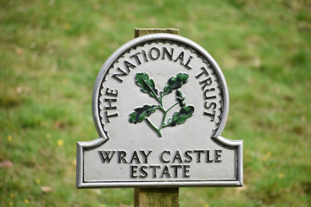 A National Trust sign with an oak leaf emblem reads "Wray Castle Estate" against a grassy background.