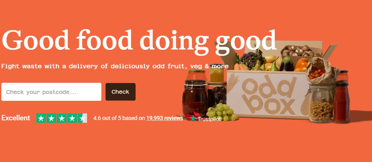 A cardboard Oddbox filled with assorted fresh fruits and vegetables is displayed on an orange background, alongside jars of preserved food.