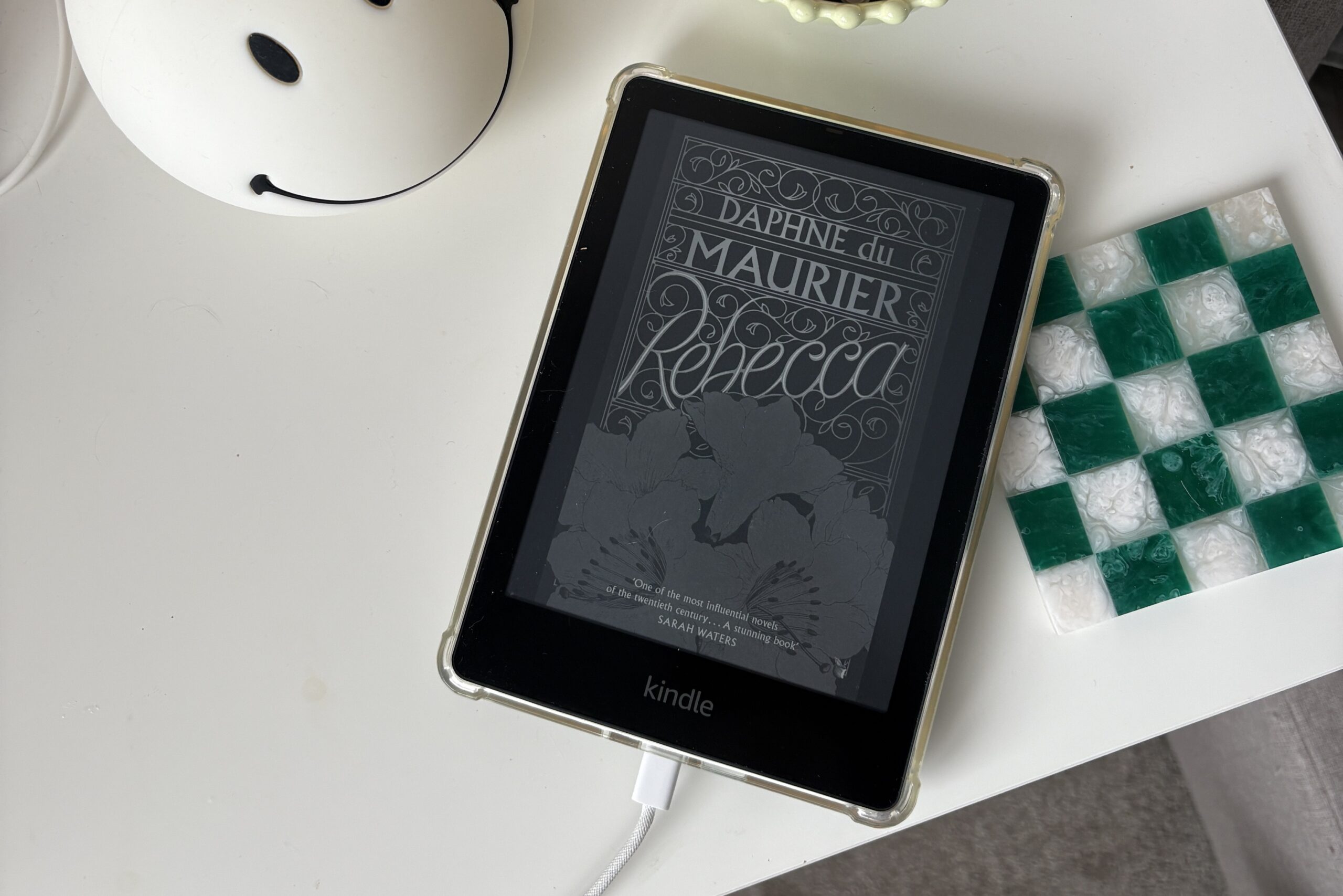 A Kindle displaying the cover of "Rebecca" by Daphne du Maurier rests on a white table next to a smiley face container and a green-and-white checkered item.