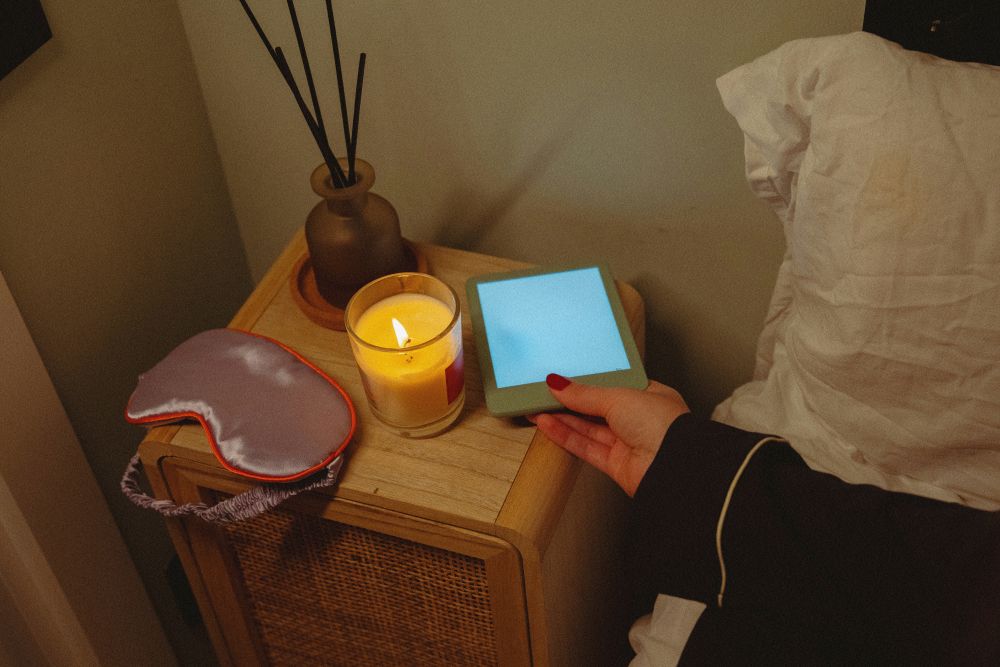 A person’s hand holds a small glowing blue device on a bedside table next to a lit candle, a reed diffuser, and a purple sleep mask.