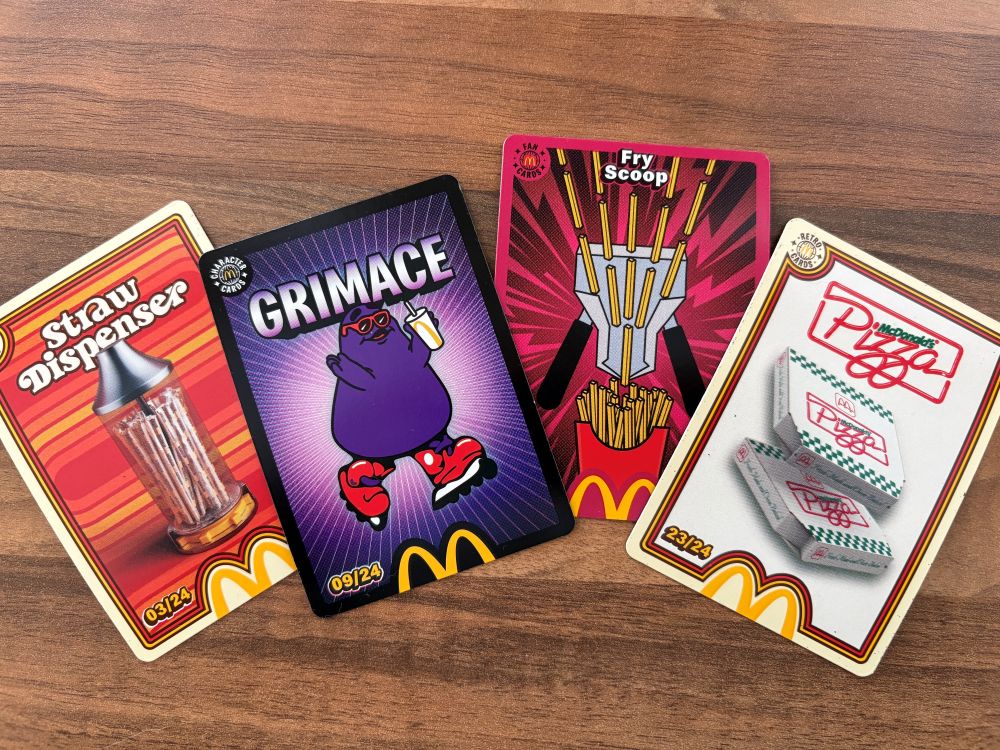 topcashback money guides mcdonalds trading cards Four McDonald’s-themed collectible cards are laid out on a wooden surface, featuring a straw dispenser, Grimace, fries, and pizza boxes.