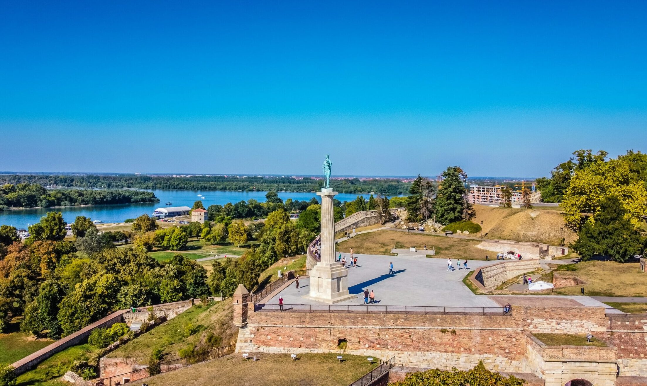 belgrade Wide view of Kalemegdan Fortress in Belgrade, Serbia, with a tall monument, green trees, people walking, and a river under a clear blue sky.