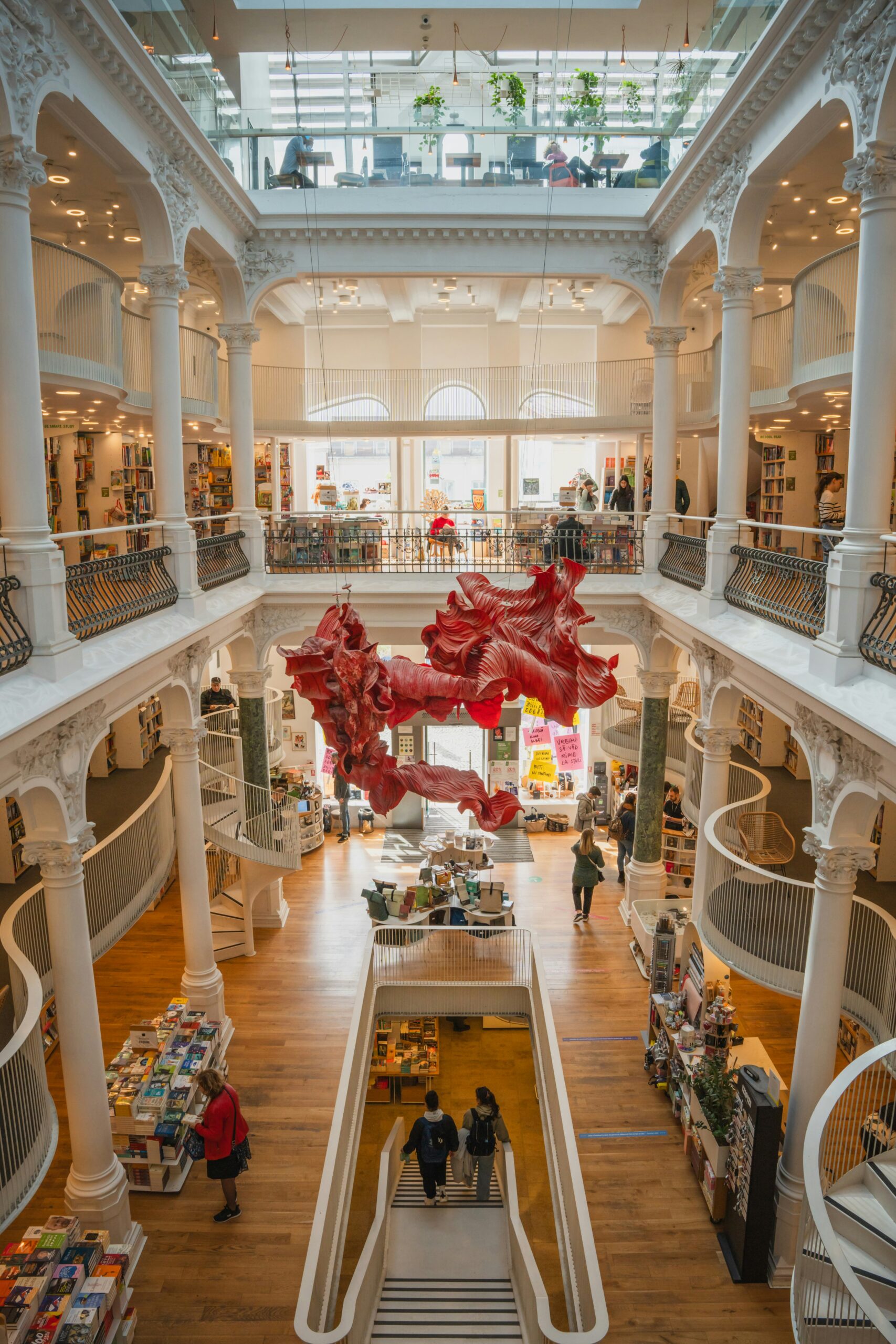 bucharest bookshop mobile Multi-level bookshop with white columns, curved staircases, wooden floors, bookshelves, and large red abstract hanging art in the centre. People are browsing and walking inside.