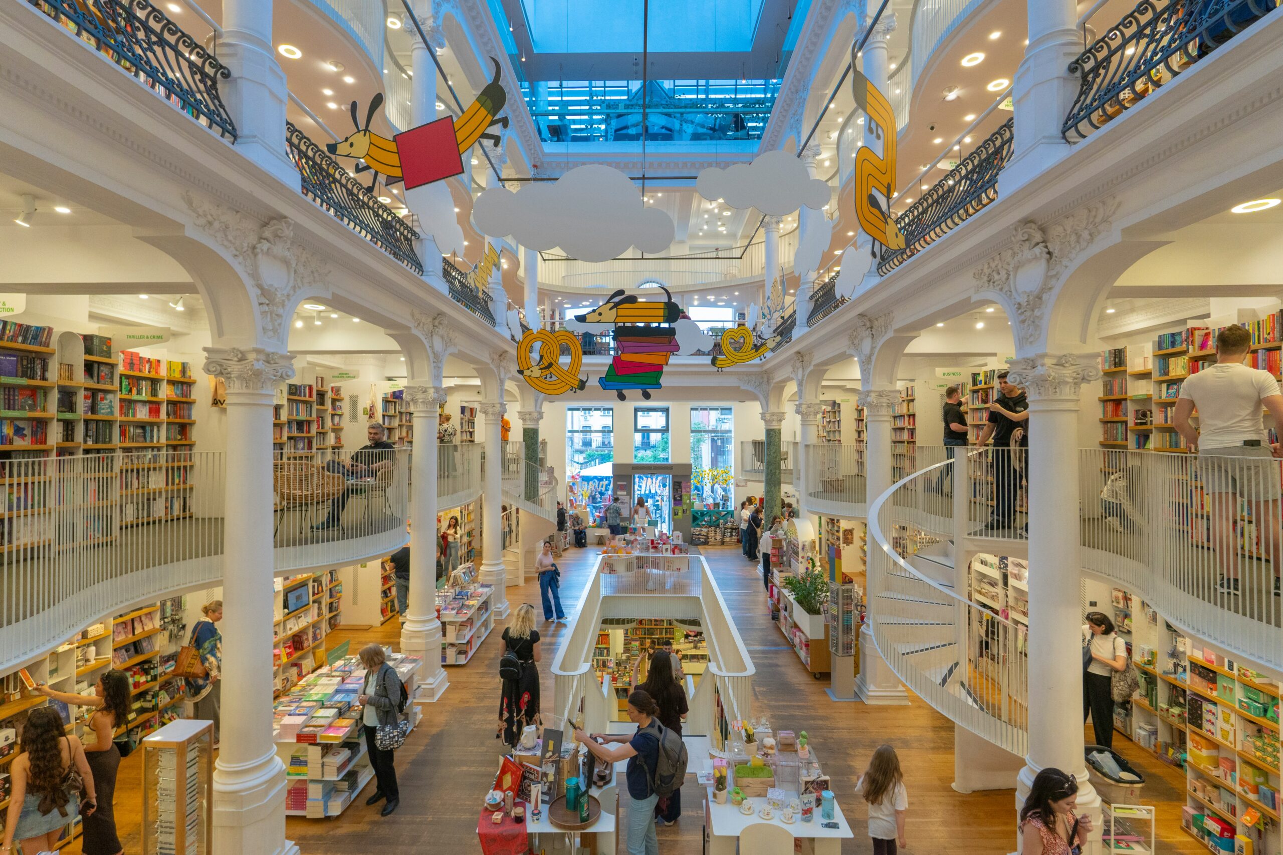bucharest bookshop A spacious, multi-level bookshop with tall white columns, spiral staircases, colourful hanging decorations, and people browsing books on shelves and tables.