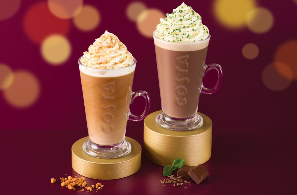 costa christmas menu 2025 Two Costa glass mugs filled with whipped cream-topped Costa drinks sit on gold stands, with caramel pieces beside one and chocolate pieces and mint leaves beside the other, against a blurred background.