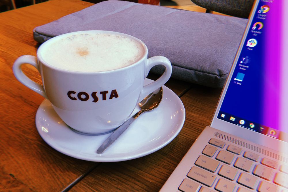 costa coffee drink A large Costa coffee cup filled with a frothy drink sits on a saucer with a spoon, next to a closed laptop and a grey case on a wooden table.