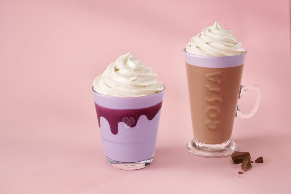 costa spring menu 2026 Two Costa drinks with whipped cream toppings in clear glasses; one is purple with dark purple syrup, the other is light brown mocha. Both are on a pink surface with small chocolate pieces nearby.