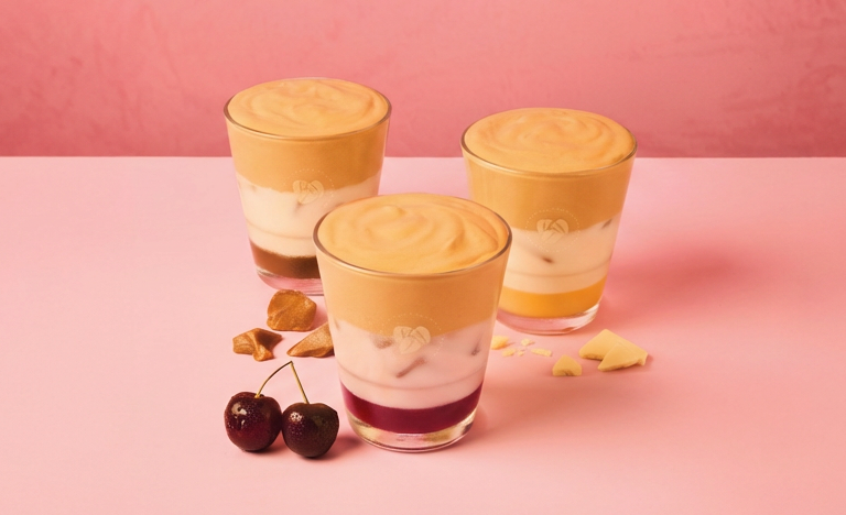 costa summer menu 2026 Three layered Costa drinks in clear glasses are displayed on a pink surface with cherries and white chocolate pieces nearby against a pink background.