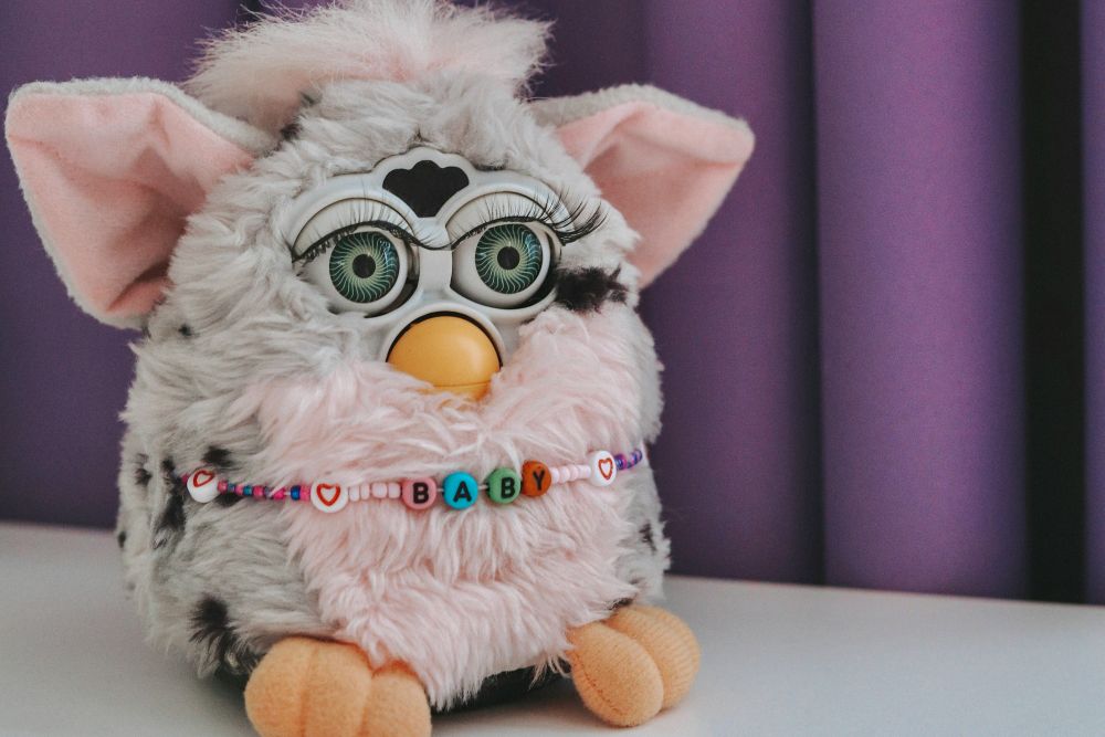 furby A gray and pink Furby toy with big eyes sits on a white surface, wearing a colorful beaded necklace spelling "BABY," in front of a purple background.