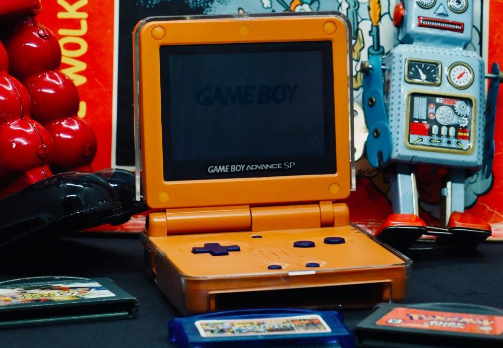 game boy advance sp An orange Game Boy Advance SP is open on a table, surrounded by game cartridges, a vintage toy robot, and other retro items.